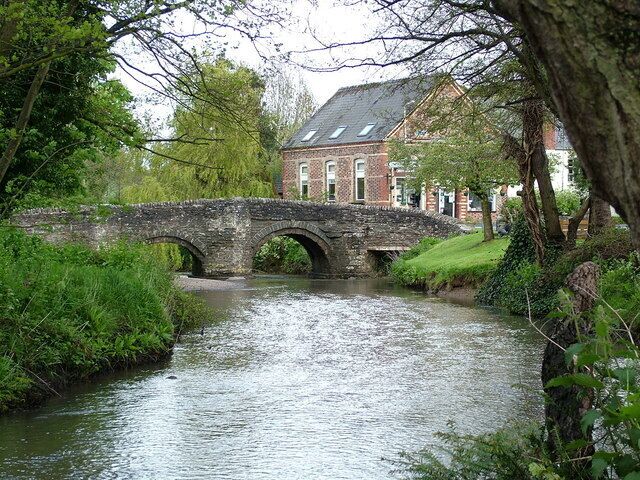 A stone bridge over a river, surrounded by greenery and a brick building.