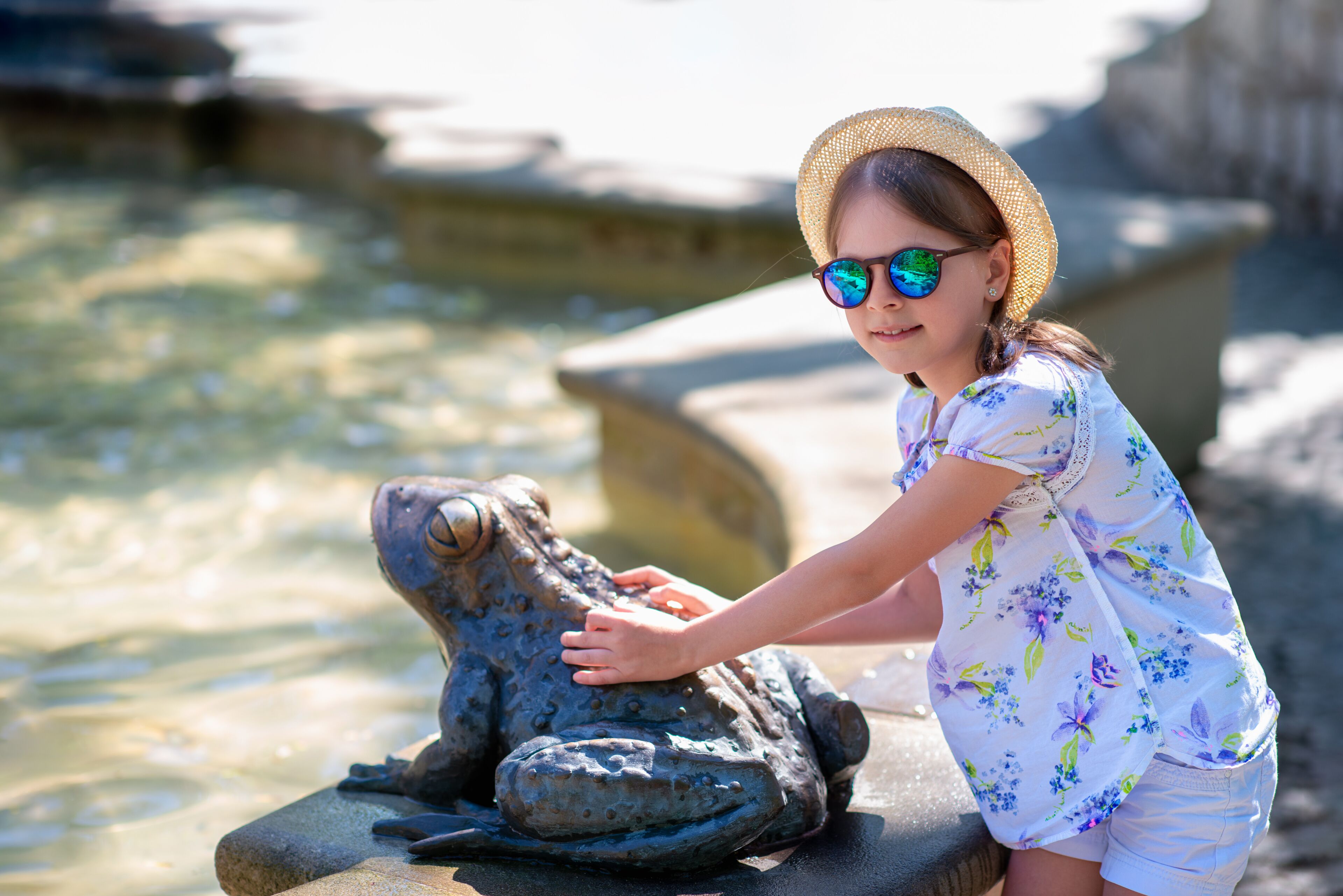 Girl in straw hat and floral shirt touching whimsical bronze frog statue by outdoor fountain in summer park, travel sightseeing and happy vacation moment, Swieradow-Zdroj, Poland