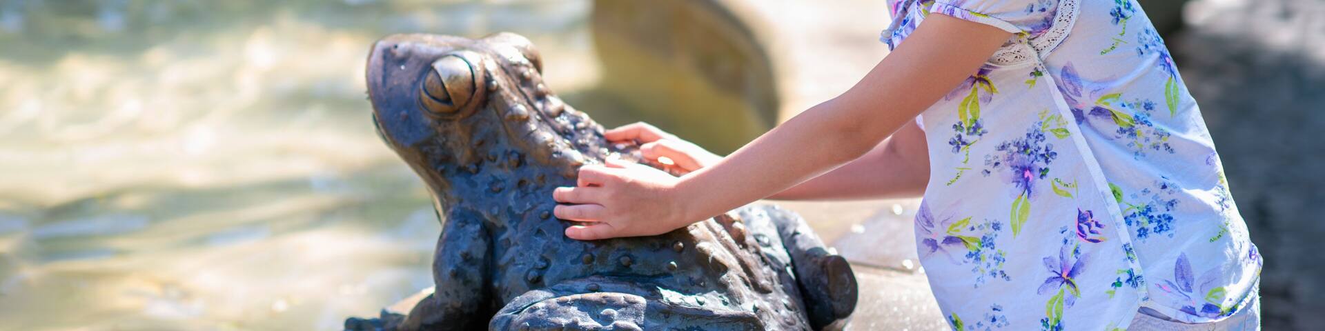 A person interacting with a frog statue by a water feature.