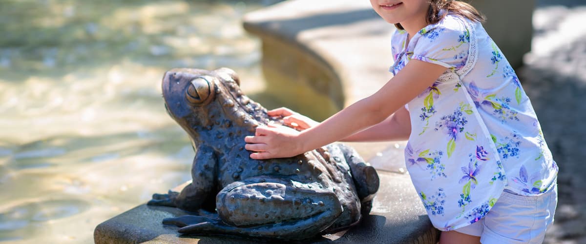A person interacting with a frog statue by a water feature.