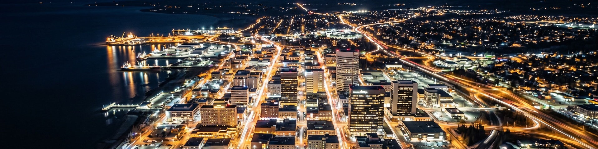 Uma cidade à noite com um rio, ruas iluminadas e montanhas nevadas ao fundo.