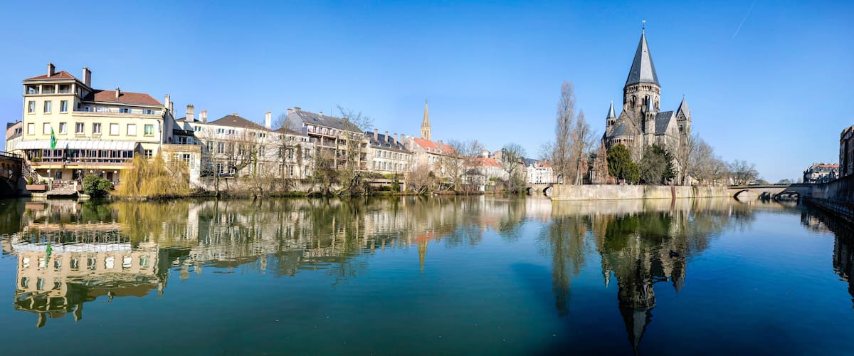 A calm river reflecting buildings, a church with a tall spire, and a bridge in a European town.