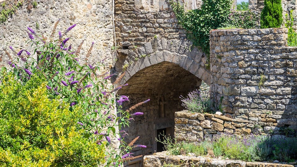 A stone pathway leading through an arched stone gateway surrounded by greenery and tall cypress trees.
