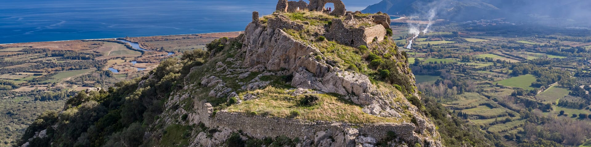 A coastal landscape with ruins on a hill overlooking the sea and distant mountains.