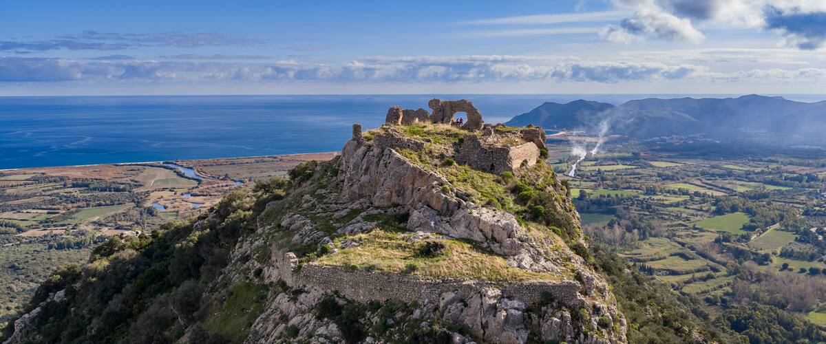 A coastal landscape with ruins on a hill overlooking the sea and distant mountains.