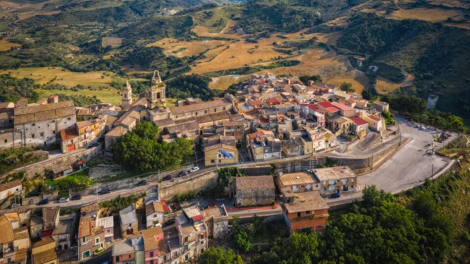 An aerial view of a historic town with a prominent church, terraced fields, and a winding road.
