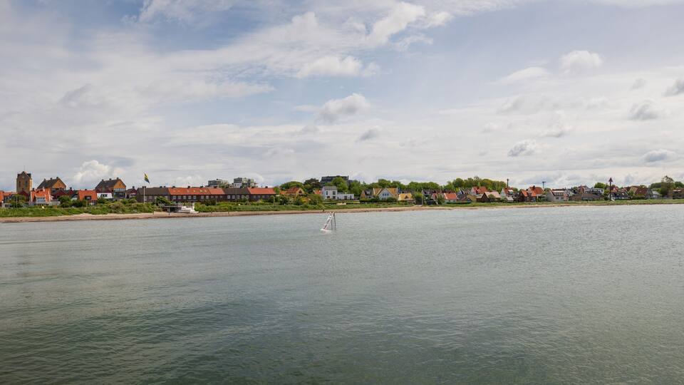 A calm river with a wooden pier on the left, a small sailboat in the water, and a row of houses and buildings along the riverbank under a partly cloudy sky.