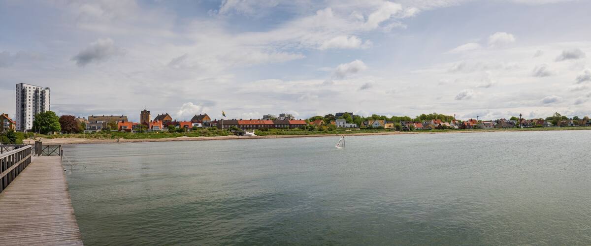 A calm river with a wooden pier on the left, a small sailboat in the water, and a row of houses and buildings along the riverbank under a partly cloudy sky.