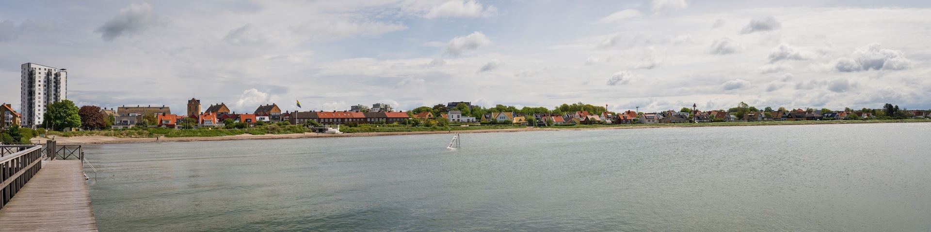 A calm river with a wooden pier on the left, a small sailboat in the water, and a row of houses and buildings along the riverbank under a partly cloudy sky.