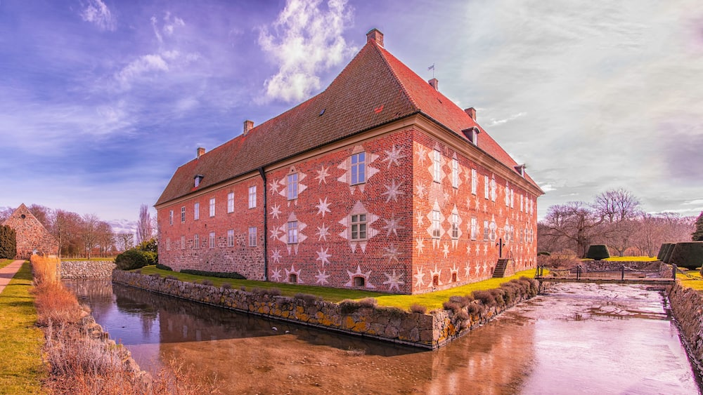 A large brick building with a red roof and star-shaped window patterns, surrounded by a moat and greenery.