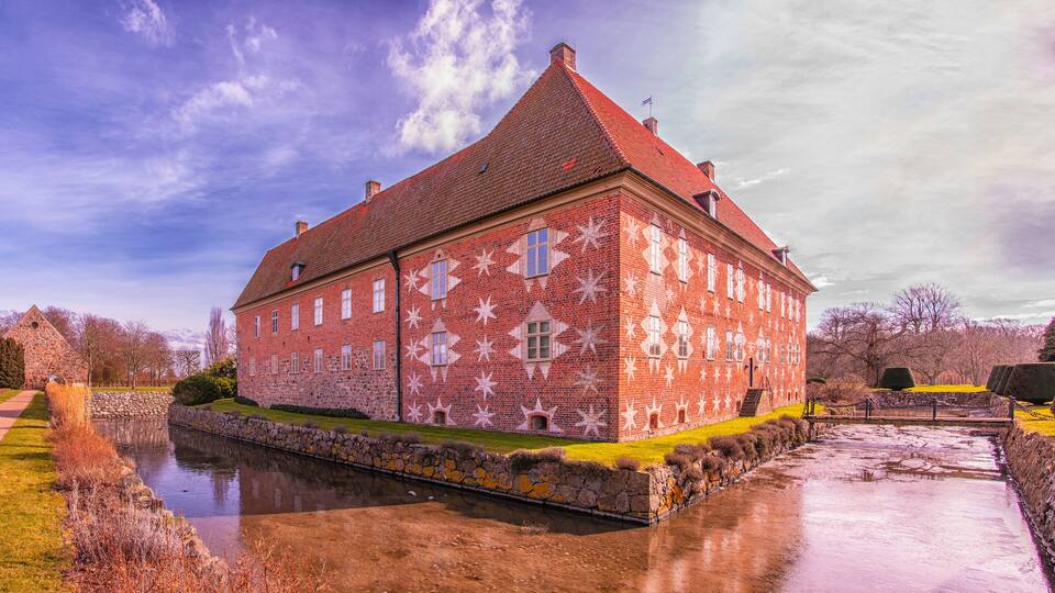 A large brick building with a red roof and star-shaped window patterns, surrounded by a moat and greenery.
