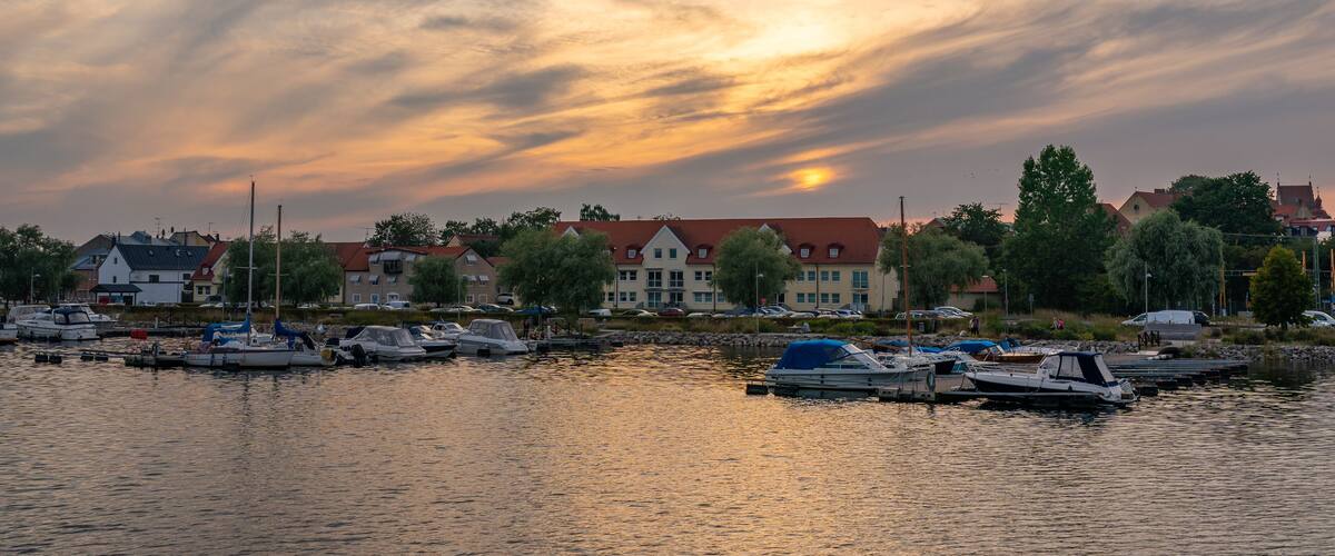 A marina with boats docked, a sunset over the water, and buildings in the background.