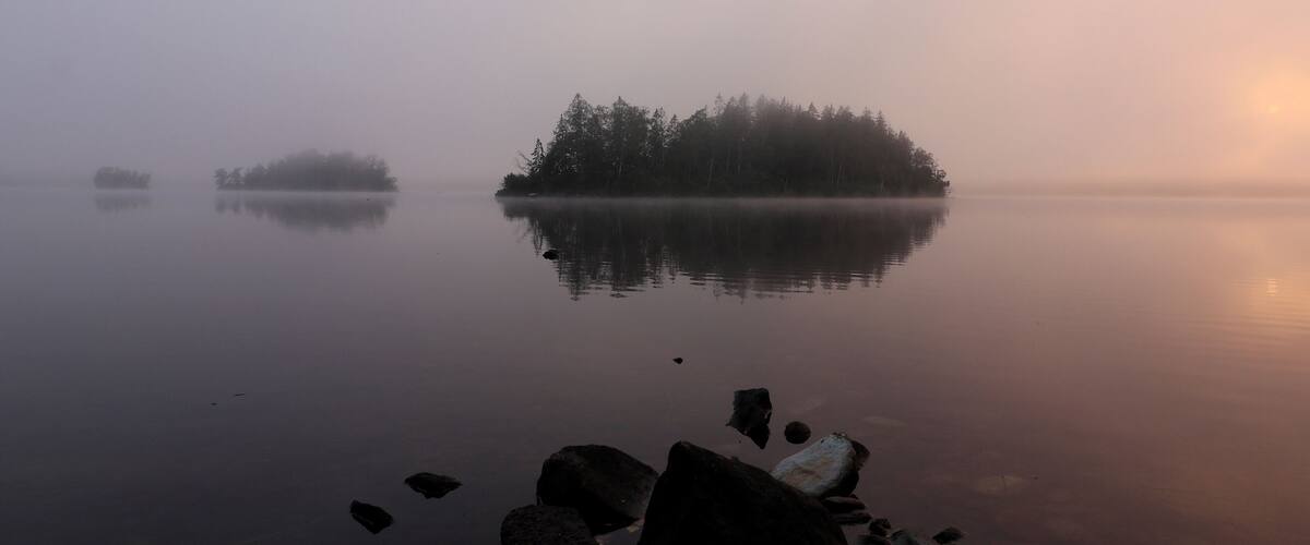 A misty lake with calm water, a small island with trees, and rocks in the foreground.
