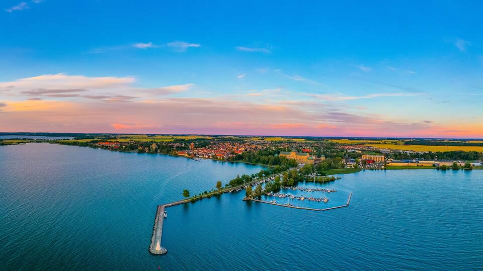 A panoramic view of a lake with a peninsula featuring a marina, a castle, and surrounding fields.