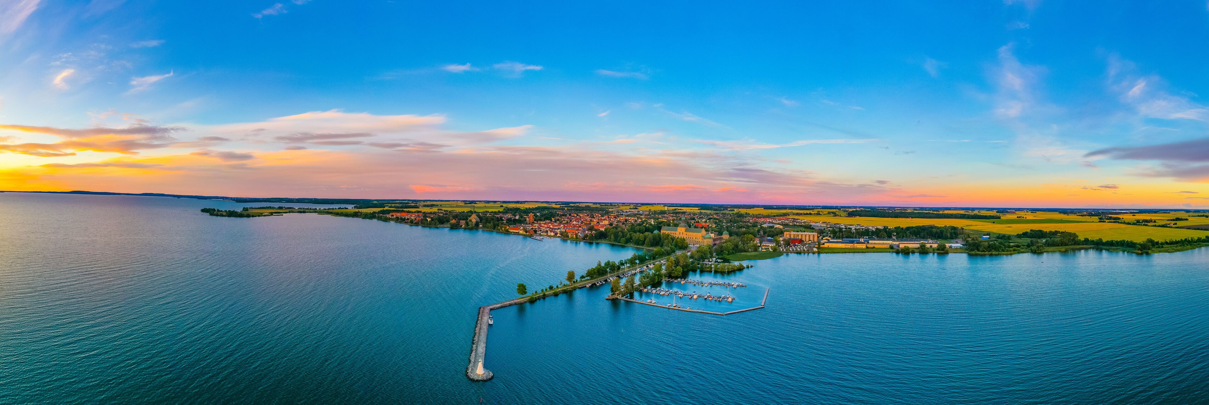 A panoramic view of a lake with a peninsula featuring a marina, a castle, and surrounding fields.