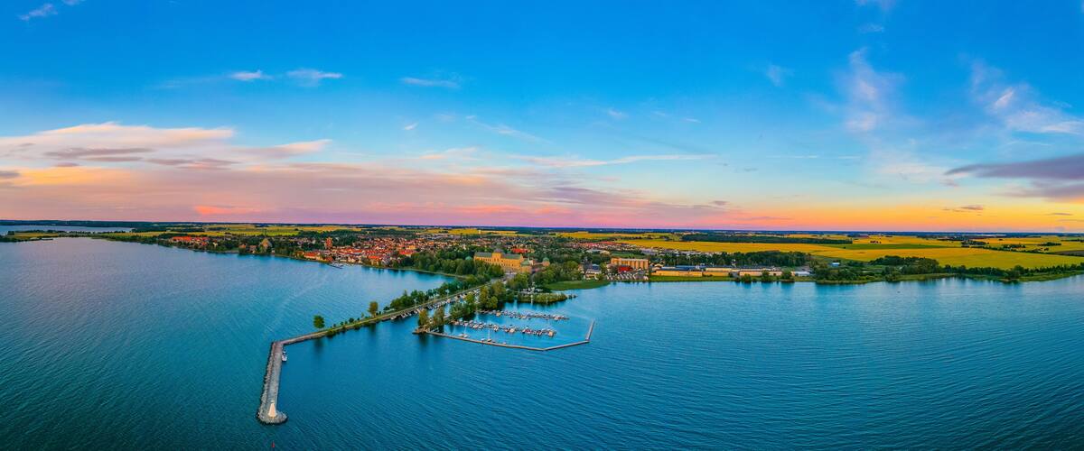 A panoramic view of a lake with a peninsula featuring a marina, a castle, and surrounding fields.