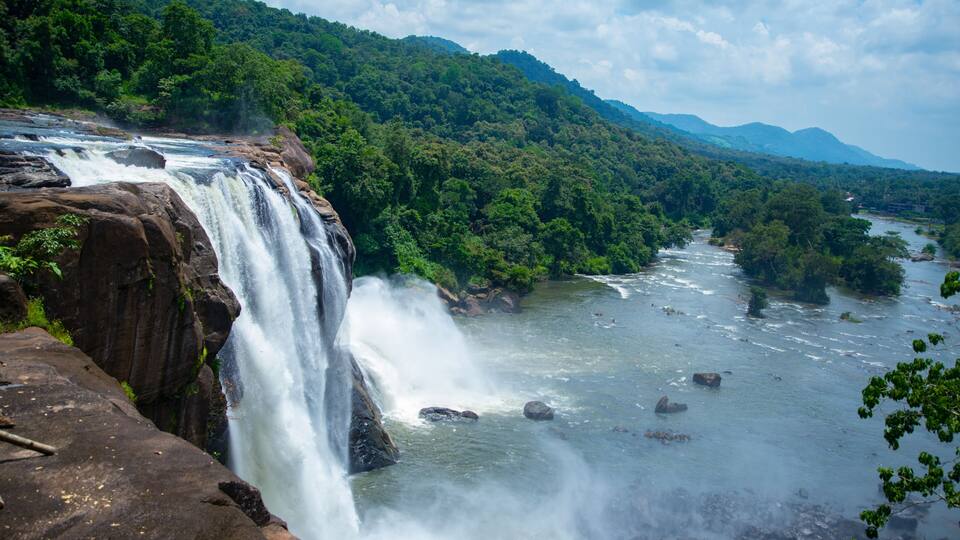 A waterfall cascading over a rocky cliff into a river surrounded by lush greenery and hills.