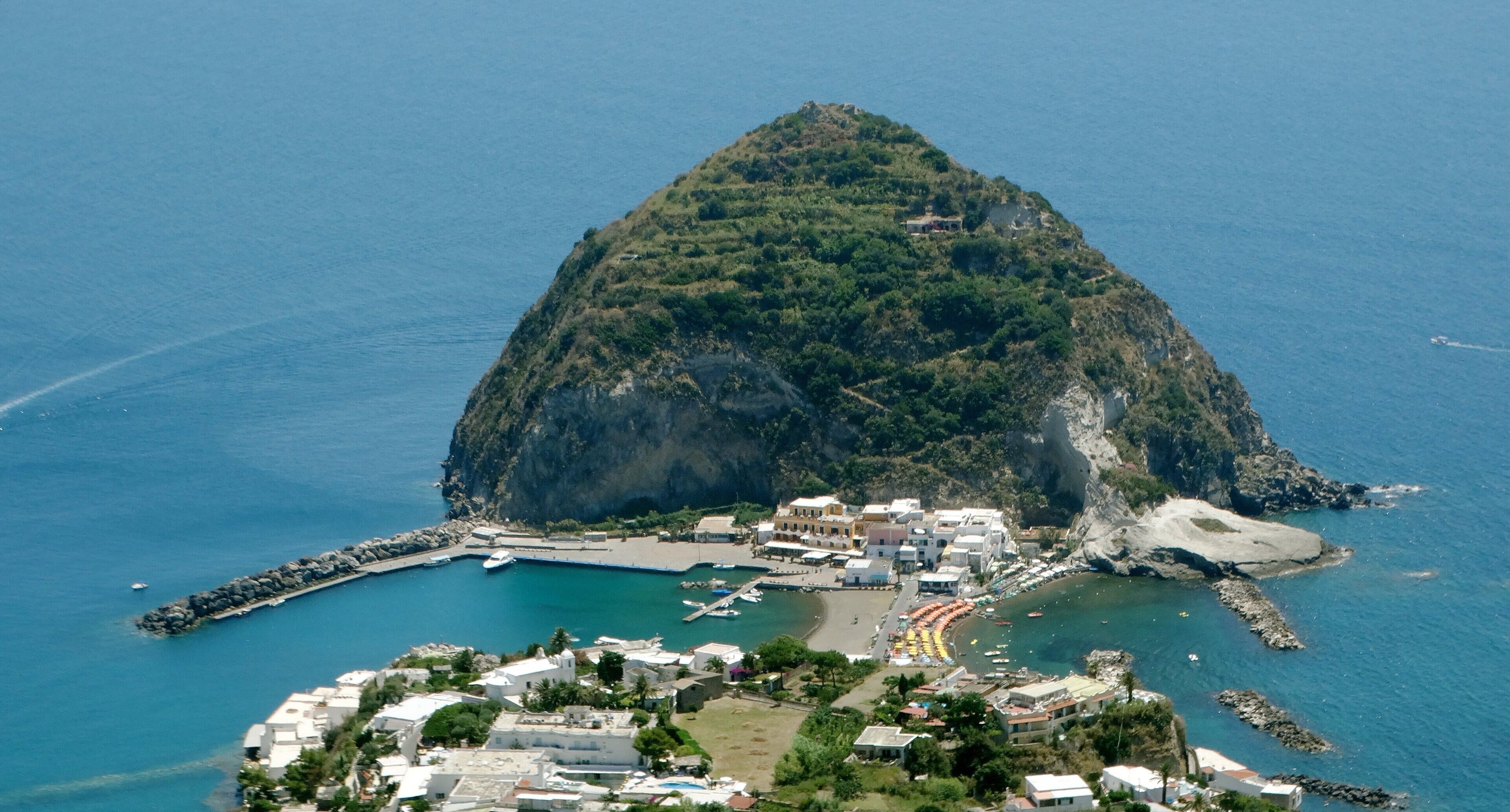 An aerial view of a coastal town with a prominent hill, a marina, and a beach area.
