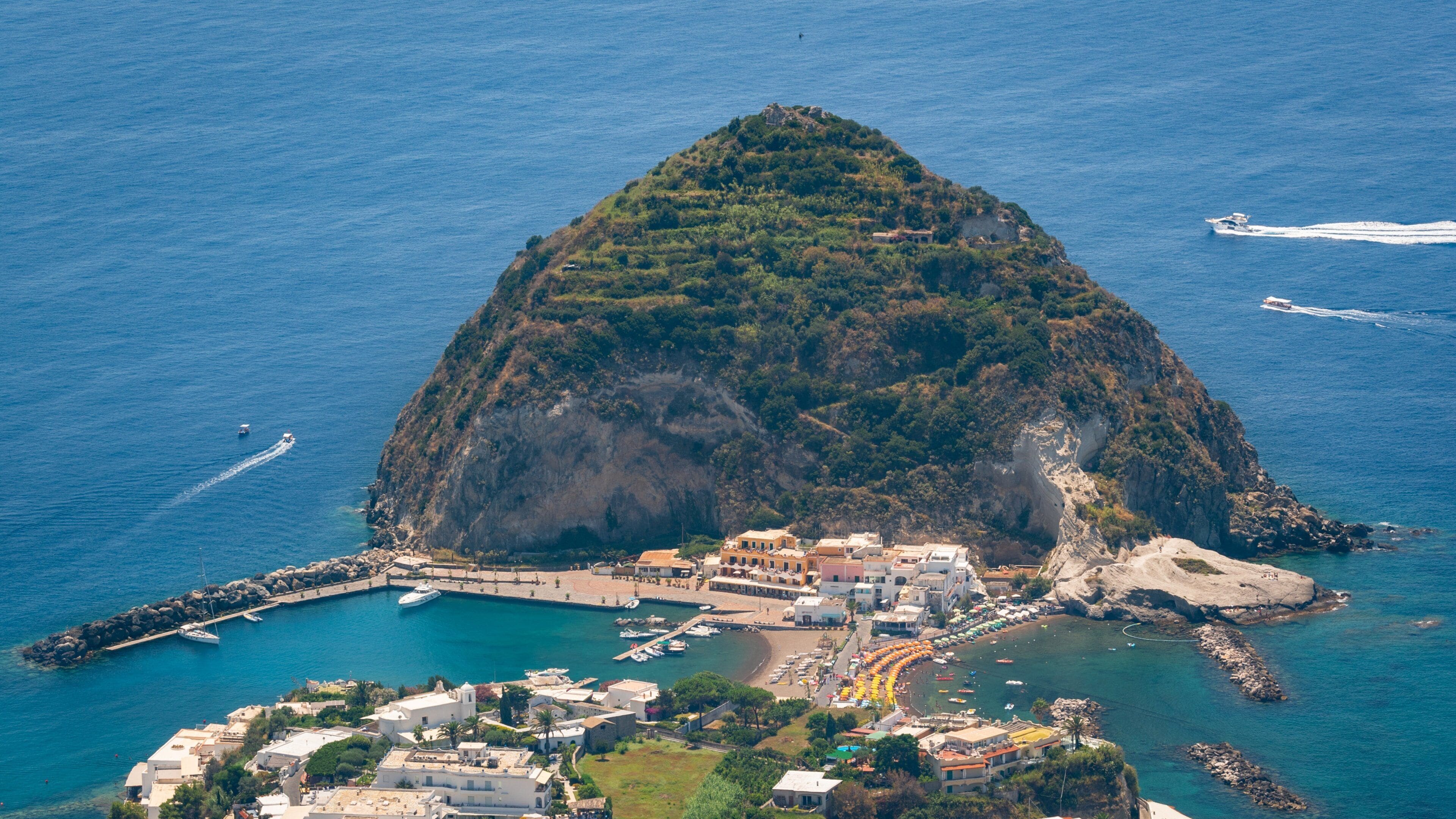 An aerial view of a small island with a dense forest, a harbor with boats, and a coastal town with buildings.