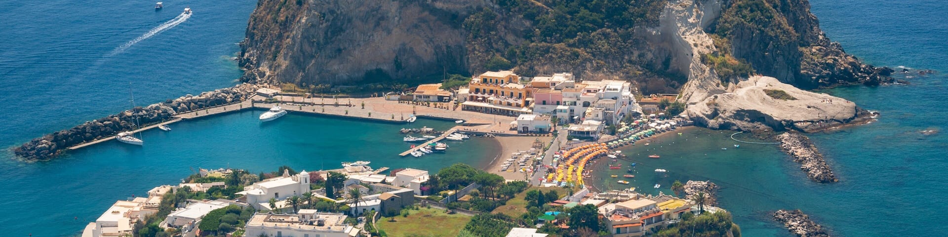 An aerial view of a small island with a dense forest, a harbor with boats, and a coastal town with buildings.