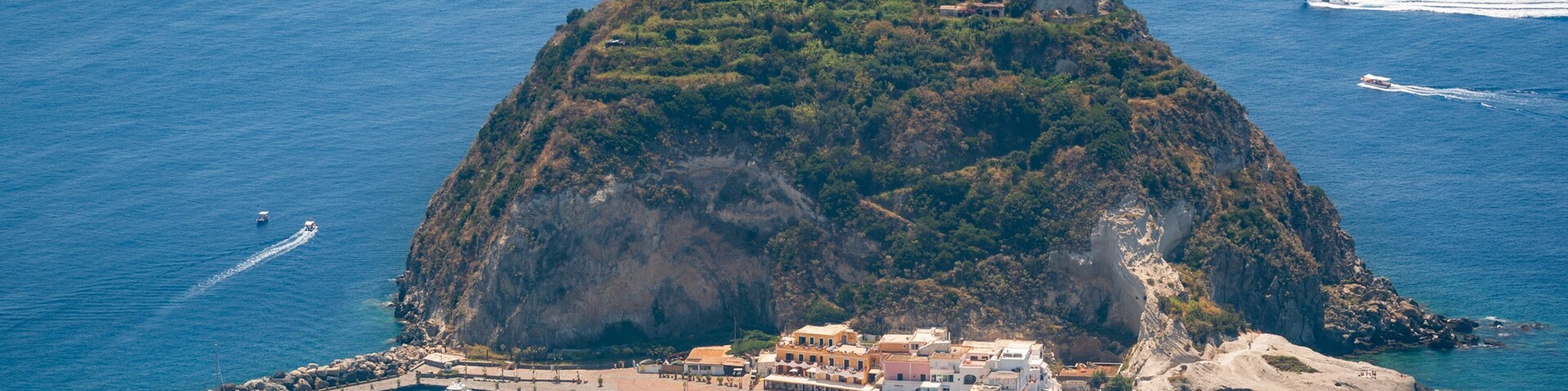 Vista aérea de una pequeña isla con un bosque tupido, un puerto con barcos y un pueblo costero con edificios.