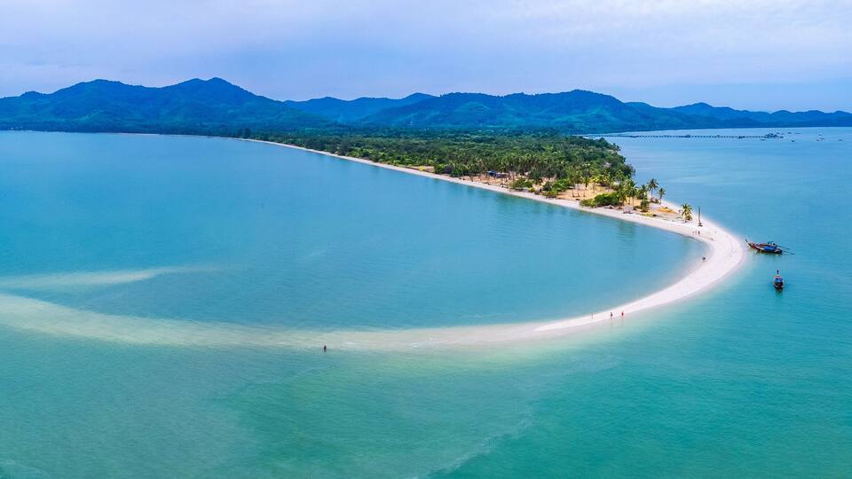 An aerial view of a coastal area with a sandy beach, clear turquoise waters, and a backdrop of green mountains.