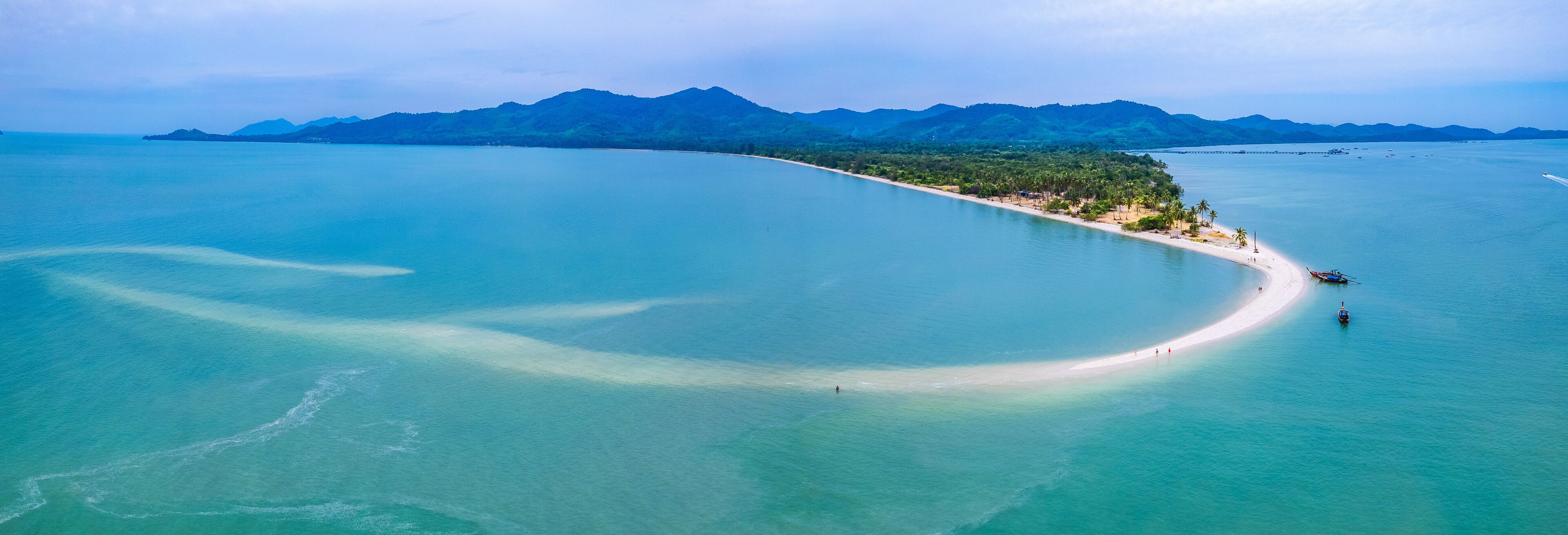 An aerial view of a coastal area with a sandy beach, clear turquoise waters, and a backdrop of green mountains.