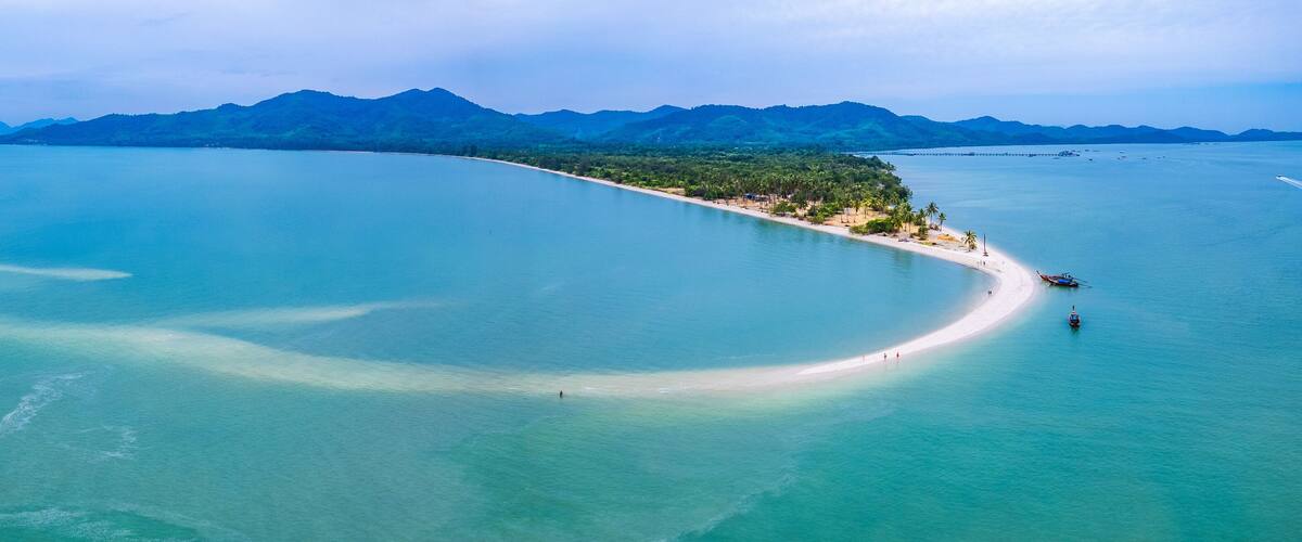 An aerial view of a coastal area with a sandy beach, clear turquoise waters, and a backdrop of green mountains.