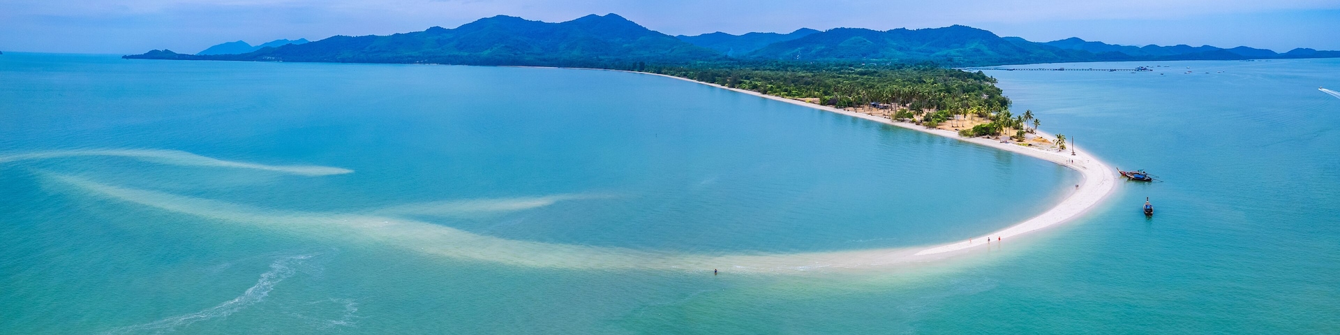An aerial view of a coastal area with a sandy beach, clear turquoise waters, and a backdrop of green mountains.