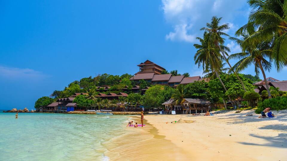 A tropical beach with clear water, palm trees, and a resort with multiple buildings.