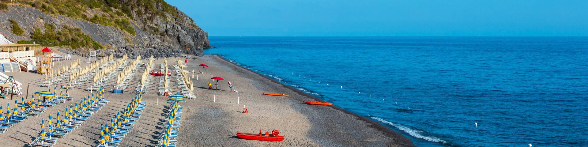 A beach with rows of beach chairs, a red boat, and a castle on a hill.