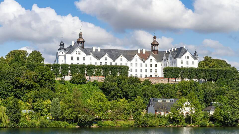A large white building with two towers, surrounded by lush greenery and a body of water.