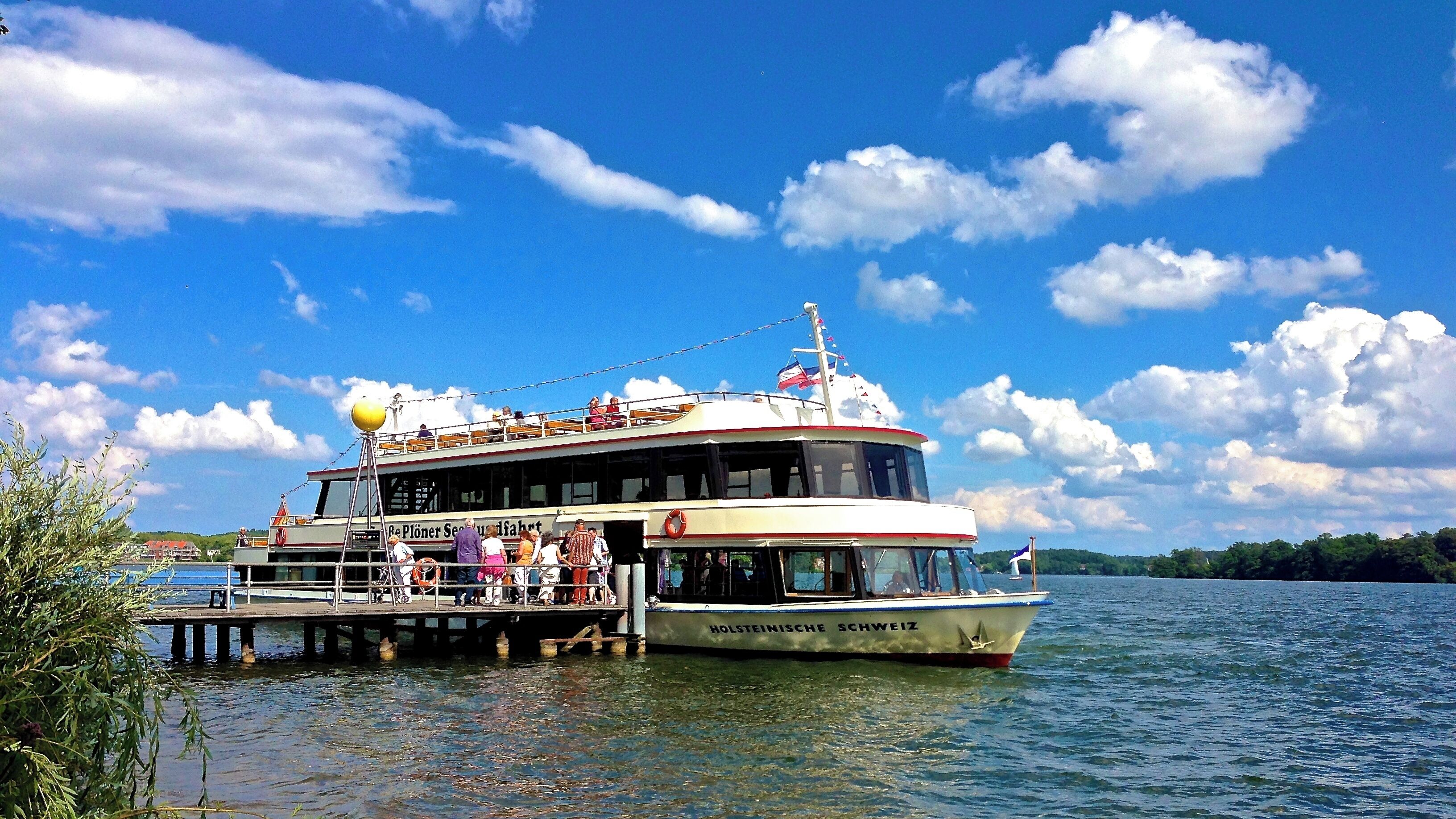 A large boat named "Holsteinische Schweiz" docked at a pier with people on board and a clear blue sky with clouds.