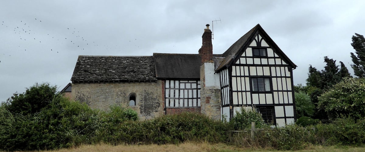 A historic half-timbered house with a stone chimney and a thatched roof, surrounded by a field and dense shrubbery.