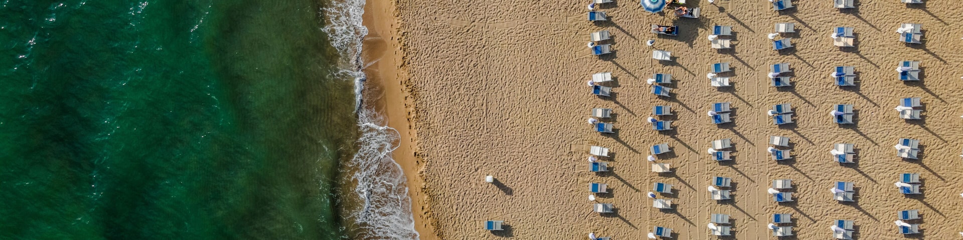 Vista aérea de uma praia com fileiras de espreguiçadeiras e guarda-sóis, e ondas do oceano quebrando suavemente na costa.