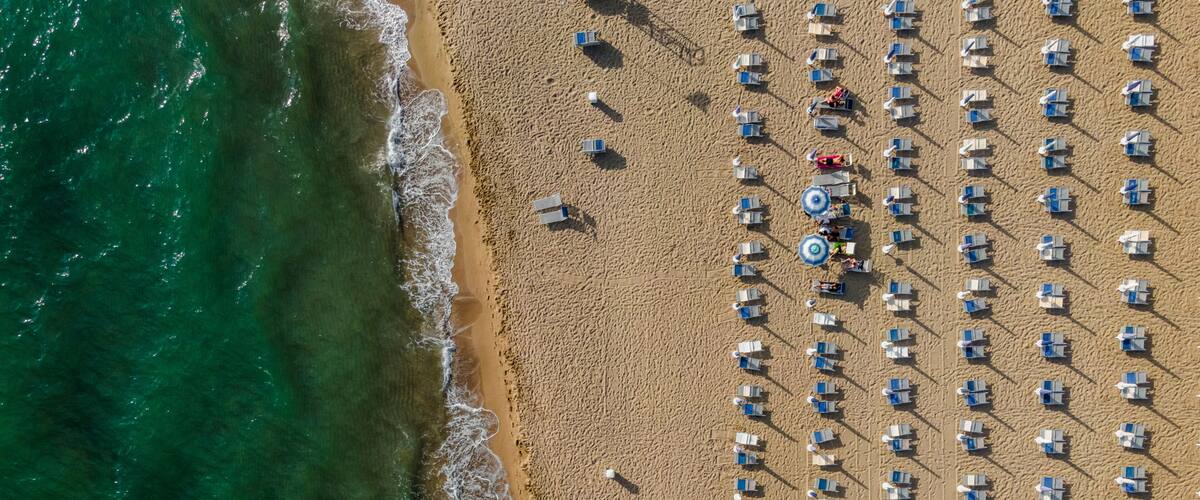 An aerial view of a beach with rows of sun loungers and umbrellas, and the ocean waves gently crashing onto the shore.