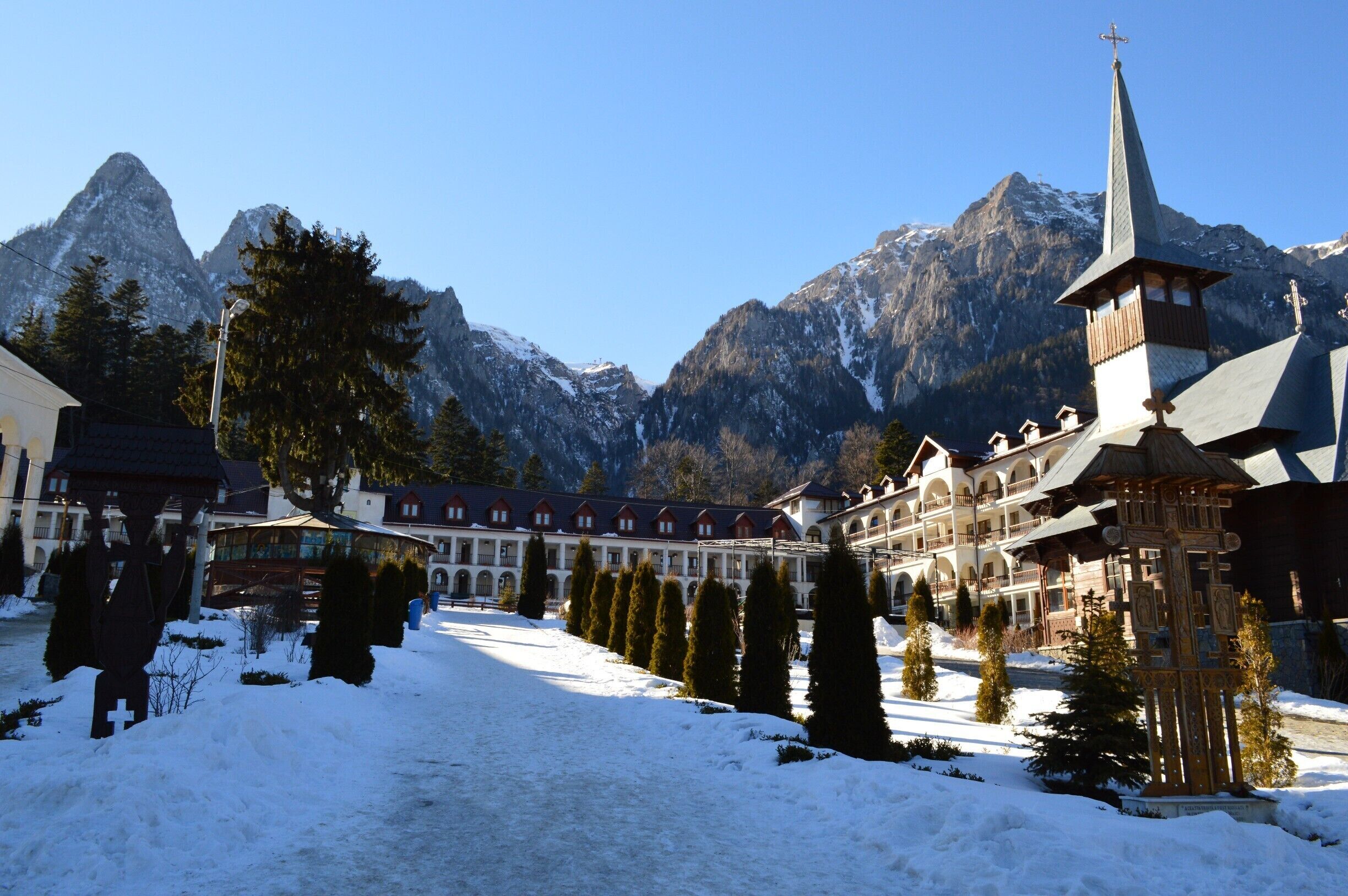 Caraiman Monastery is an orthodox monastery, which is situated in town Buşteni, Prahova district, at the foot of the massif Caraiman. The Cross from above the Caraiman seems to guard the path, which leeds the steps of the pilgrims towards the monastery. Going towards the cab from Buşteni, at a time, on the left side, there are an indicator and an icon showing the path with stone tiles in patches, path leading towards the monastery.
The secretly like walking path, under the dark crowns of the fir trees is eventually reaching an oasis of light, the massif Caraiman emerges in all its splendour. In this glade, Father Gherontie Puiu built, by the grace of Lord and with Virgin’s Mary help, a great monastery. 
Buşteni is a small mountain town situated in the northern part of Prahova district, Muntenia, located in the center of Romania. It is situated on the Prahova Valley, at he foot of the Bucegi Mountains, which have the maximum altitude of 2.505 m. Situated at a distance of 135 km from Bucharest, it stretches about 7 km along the Prahova Valley, between the “Deer Ford” and the tunnel dug into the mountain spur “Long Edge”.
The average height of the town Buşteni is of 850 m. It is one of the most popular mountain resorts, offering spectacular views, a lot of opportunities and holiday activities from skiing to hiking. It’s got a population of 10.463 inhabitants.
The Caraiman Monastery is situated at a height of about 900 m, being one of the highest altitudes where a place of worship has ever been built in our country. 