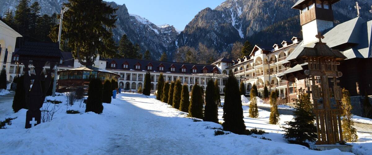 Caraiman Monastery is an orthodox monastery, which is situated in town Buşteni, Prahova district, at the foot of the massif Caraiman. The Cross from above the Caraiman seems to guard the path, which leeds the steps of the pilgrims towards the monastery. Going towards the cab from Buşteni, at a time, on the left side, there are an indicator and an icon showing the path with stone tiles in patches, path leading towards the monastery.
The secretly like walking path, under the dark crowns of the fir trees is eventually reaching an oasis of light, the massif Caraiman emerges in all its splendour. In this glade, Father Gherontie Puiu built, by the grace of Lord and with Virgin’s Mary help, a great monastery.
Buşteni is a small mountain town situated in the northern part of Prahova district, Muntenia, located in the center of Romania. It is situated on the Prahova Valley, at he foot of the Bucegi Mountains, which have the maximum altitude of 2.505 m. Situated at a distance of 135 km from Bucharest, it stretches about 7 km along the Prahova Valley, between the “Deer Ford” and the tunnel dug into the mountain spur “Long Edge”.
The average height of the town Buşteni is of 850 m. It is one of the most popular mountain resorts, offering spectacular views, a lot of opportunities and holiday activities from skiing to hiking. It’s got a population of 10.463 inhabitants.
The Caraiman Monastery is situated at a height of about 900 m, being one of the highest altitudes where a place of worship has ever been built in our country.