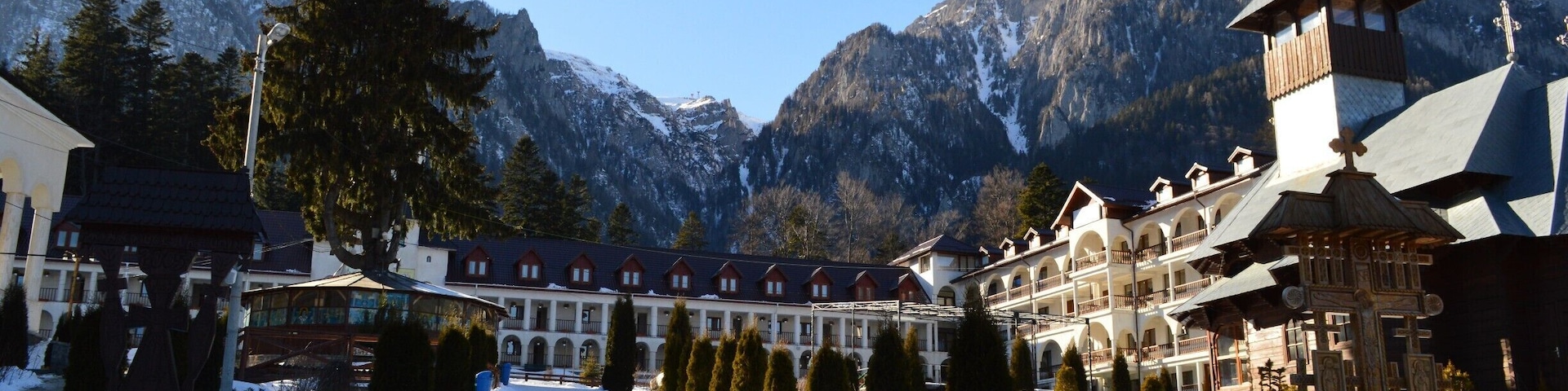 Caraiman Monastery is an orthodox monastery, which is situated in town Buşteni, Prahova district, at the foot of the massif Caraiman. The Cross from above the Caraiman seems to guard the path, which leeds the steps of the pilgrims towards the monastery. Going towards the cab from Buşteni, at a time, on the left side, there are an indicator and an icon showing the path with stone tiles in patches, path leading towards the monastery.
The secretly like walking path, under the dark crowns of the fir trees is eventually reaching an oasis of light, the massif Caraiman emerges in all its splendour. In this glade, Father Gherontie Puiu built, by the grace of Lord and with Virgin’s Mary help, a great monastery.
Buşteni is a small mountain town situated in the northern part of Prahova district, Muntenia, located in the center of Romania. It is situated on the Prahova Valley, at he foot of the Bucegi Mountains, which have the maximum altitude of 2.505 m. Situated at a distance of 135 km from Bucharest, it stretches about 7 km along the Prahova Valley, between the “Deer Ford” and the tunnel dug into the mountain spur “Long Edge”.
The average height of the town Buşteni is of 850 m. It is one of the most popular mountain resorts, offering spectacular views, a lot of opportunities and holiday activities from skiing to hiking. It’s got a population of 10.463 inhabitants.
The Caraiman Monastery is situated at a height of about 900 m, being one of the highest altitudes where a place of worship has ever been built in our country.