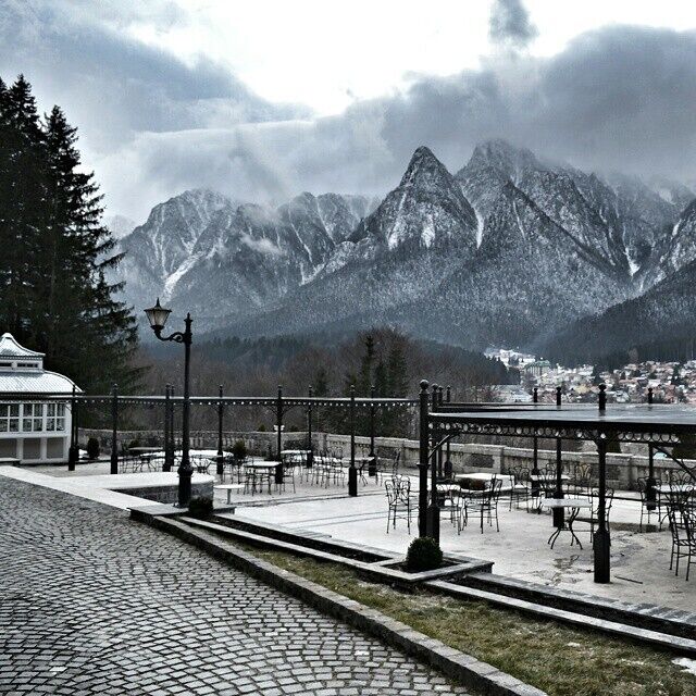 A cobblestone pathway leading to a gazebo with tables and chairs, overlooking snow-capped mountains and a town in the valley below.