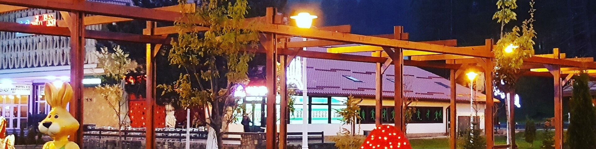 Un parque con una gran estatua de conejo, una escultura luminosa en forma de huevo rojo y amarillo, y un pérgola de madera.