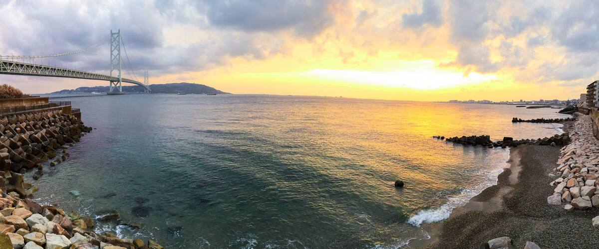 A coastal sunset with a bridge, rocky shoreline, and distant cityscape.