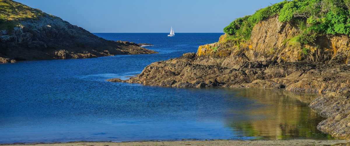 A coastal scene with a sailboat on the water, rocky cliffs, and a sandy beach.