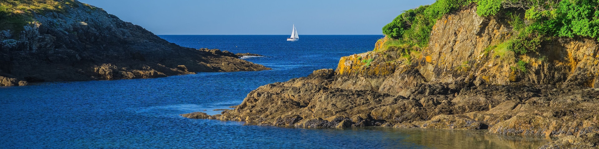 A coastal scene with a sailboat on the water, rocky cliffs, and a sandy beach.