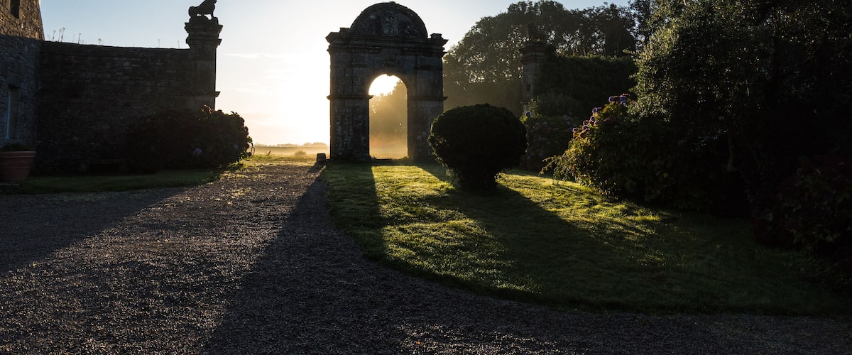 An ancient stone archway with a sun shining through it, surrounded by greenery and a gravel path.