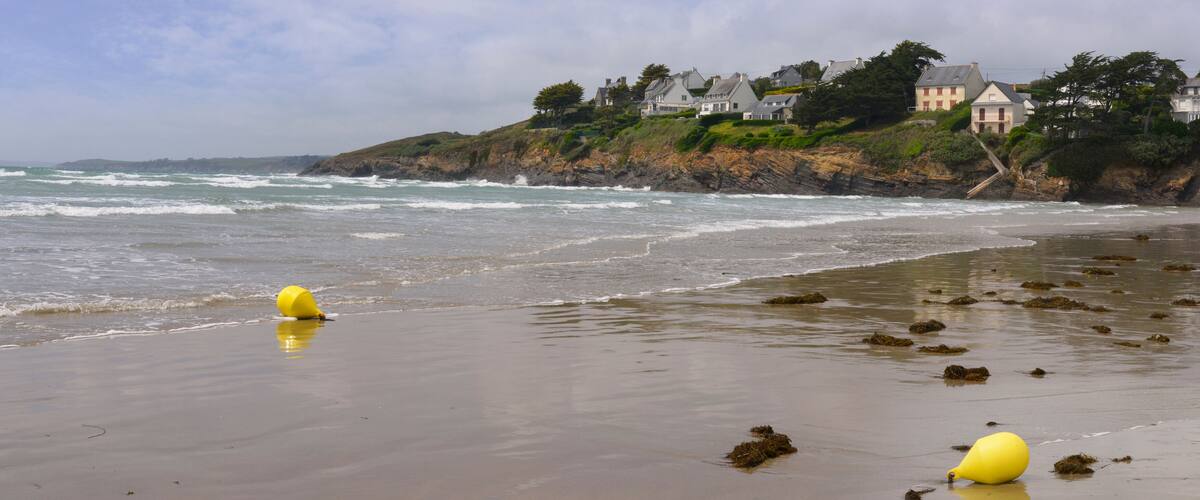 A beach with waves, yellow buoys, and houses on a cliff.