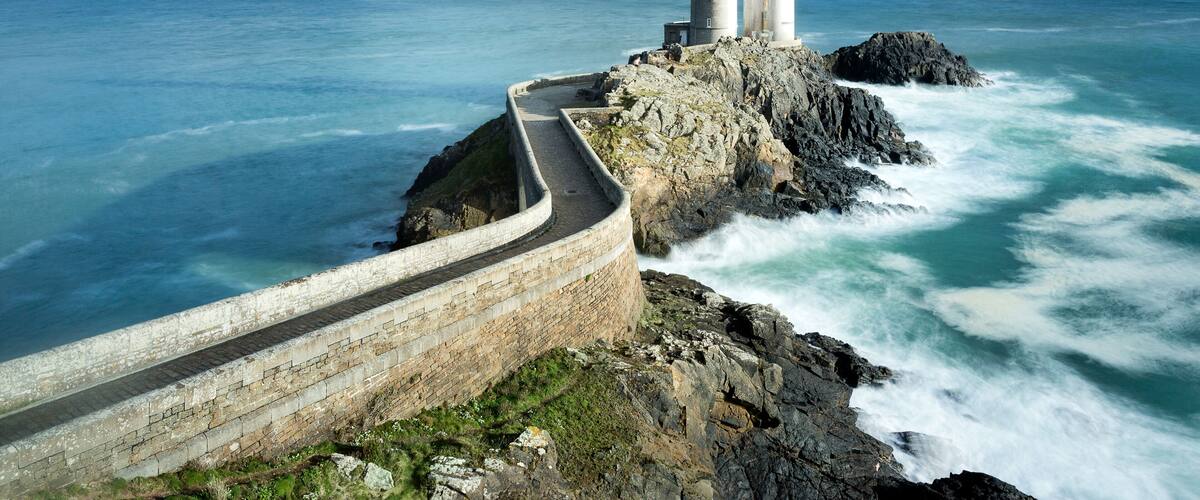 A lighthouse on a rocky coastline with a stone wall and the ocean in the background.