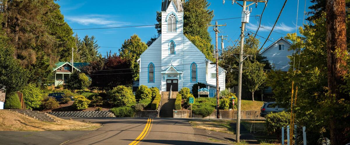 A white church with a steeple and a cross on top, surrounded by trees and a road.