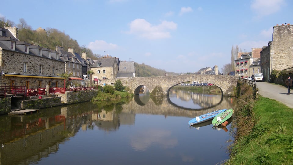 A calm river with a stone bridge, stone buildings, and a small boat docked on the riverbank.