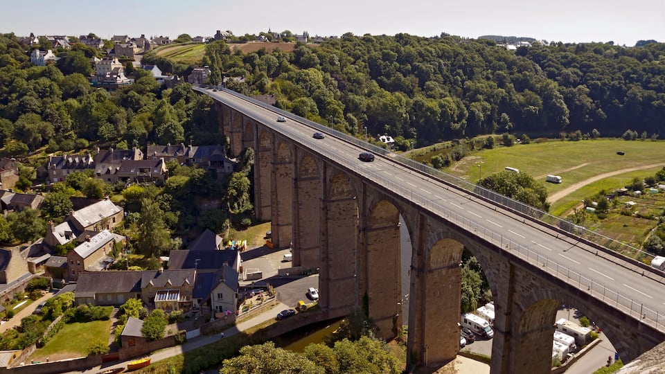 A large stone bridge spanning a river, surrounded by lush greenery and a small town.